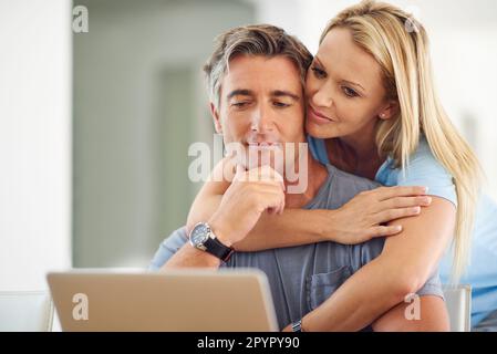 Pensi che dovrei postare questo amore. una donna matura e attraente che abbraccia il suo bel marito maturo mentre usa un computer portatile in casa. Foto Stock