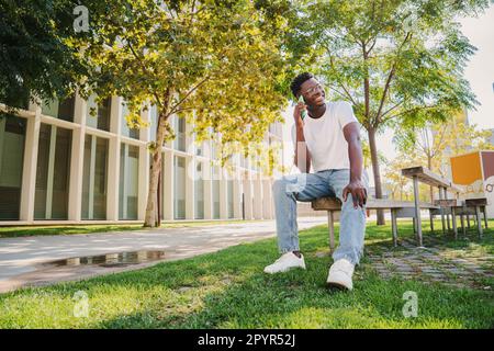 Felice studente afro americano università adolescente avere una conversazione divertente e ridere. Ritratto di un giovane che parla utilizzando un telefono cellulare seduto su una panca all'aperto nel campus universitario. Foto di alta qualità Foto Stock