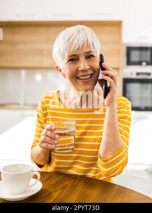 Donna piuttosto matura che parla sul telefono cellulare e tiene un bicchiere d'acqua al tavolo da pranzo a casa Foto Stock