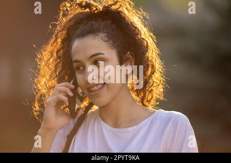 Ritratto di ragazza adolescente cute che parla sul telefono cellulare all'aperto in natura al tramonto in primavera o in estate Foto Stock