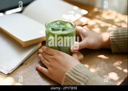 Immagine ravvicinata di una donna in un comodo maglione che tiene un bicchiere di tè verde matcha ghiacciato a un tavolo in un bel caffè minimal. Foto Stock