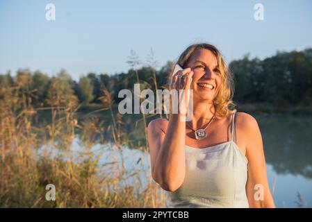 Donna allegra sorridente mentre parla con uno smartphone a Riverbank, Baviera, Germania Foto Stock