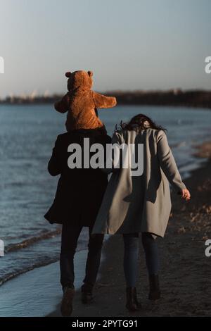 Una coppia che cammina lungo una spiaggia sabbiosa, l'uomo che porta il suo bambino indossando un costume da orsacchiotto Foto Stock