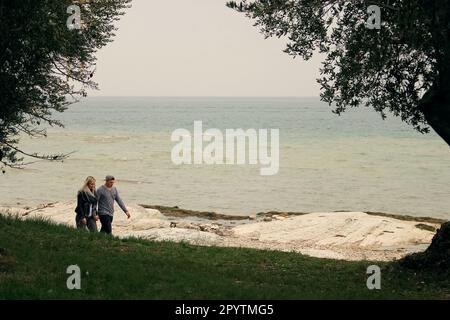 una giovane coppia tradizionale, un uomo bianco e una donna bianca, innamorati e in mano, camminano lungo la riva di un grande lago senza paura della pioggia. Foto Stock