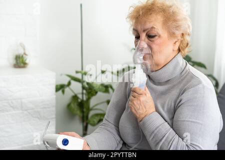 La donna anziana malata ha un'infezione virale o influenza, respirazione laboriosa, asma. Una donna anziana utilizza inalatore, nebulizzatore per il trattamento autonomo seduto Foto Stock
