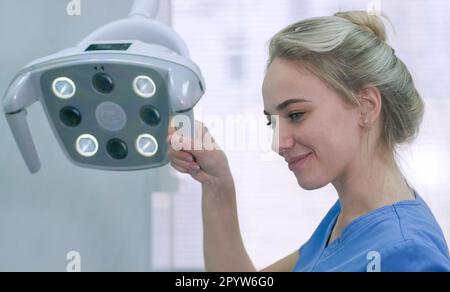 Bella infermiera che regola la luce dentale in clinica. L'assistente dentista prepara l'apparecchiatura di illuminazione prima del trattamento e sorride al paziente Foto Stock
