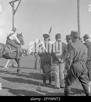 Il pilota partecipa al ring riding a Warnemünde. Nella corsa ad anello, una lancia è messa attraverso un anello appeso al ponteggio. Gli spettatori si trovano accanto al ponteggio. Un uomo indossa un'uniforme dell'esercito prussiano. Proprio nella foto è un uomo SA. Foto Stock