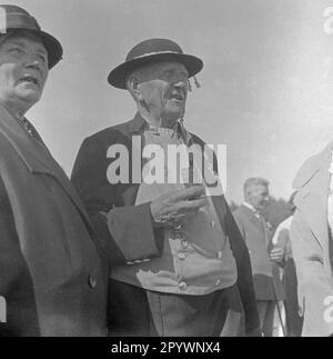 Gli spettatori guardano il ring a Warnemünde. Nella corsa ad anello, una lancia è messa attraverso un anello appeso ad un ponteggio. L'uomo indossa abiti tradizionali e fuma una pipa. Foto Stock