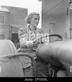 Una donna bionda che indossa un abito e un grembiule guida un trattore. Sullo sfondo, edifici cortile. Foto non datata. Foto Stock