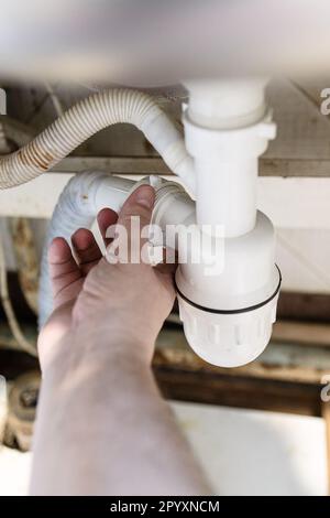 la mano dell'idraulico disassembla il sifone di plastica vecchio vicino in su nella cucina della casa del villaggio per pulire lo scarico Foto Stock