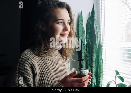 Una giovane donna serena assapora una tazza calda di tè mentre ammiri lo splendido paesaggio urbano da una finestra, prendendosi un momento tranquillo a se stessa per rilassarsi e. Foto Stock
