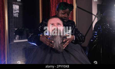 Una ragazza afro-americana del parrucchiere raddrizza la sua barba con le mani ad un cliente maschio seduto in una poltrona Foto Stock