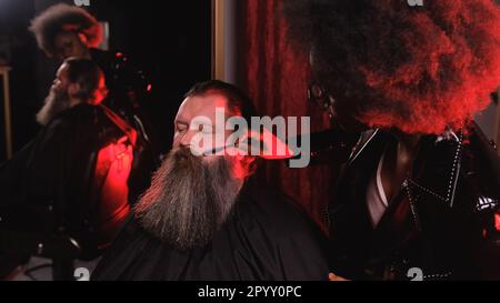 Una stilista afro-americana in un impermeabile in pelle nera pettina la barba di un cliente in un barbiere Foto Stock