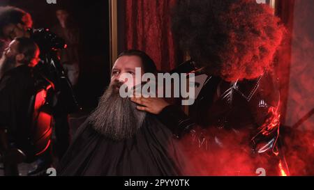 Una stilista afro-americana in un impermeabile in pelle nera pettina la barba di un cliente in un barbiere Foto Stock