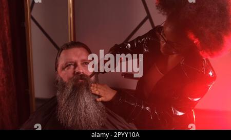 Una stilista afro-americana in un impermeabile in pelle nera pettina la barba di un cliente in un barbiere Foto Stock