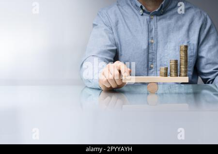 Close-up di un imprenditore la mano di bilanciamento delle monete impilate su altalena in legno con il dito sulla scrivania Foto Stock