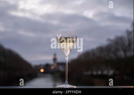 Bicchiere di vino bianco frizzante francese con champagne bolle all'aperto con vista sulle acque verdi del fiume Marne e le luci del viale Promenade in mor Foto Stock