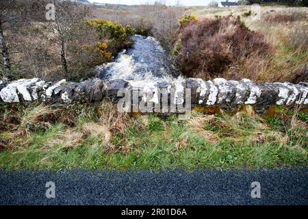 strada e vecchio ponte di pietra sul piccolo fiume torrente in alluvione nella remota regione rurale di montagna della contea donegal repubblica d'irlanda Foto Stock