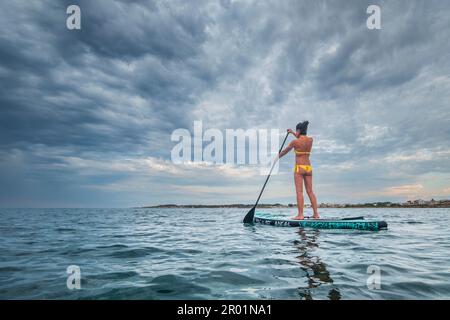Donna che paddling su una tavola da surf sotto un cielo drammatico, SA Rapita, Maiorca, Isole Baleari, Spagna. Foto Stock