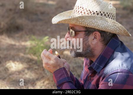 Uomo con cappello, occhiali da sole, camicia a scacchi e barba bianca che illumina una sigaretta con un accendisigari nella foresta Foto Stock