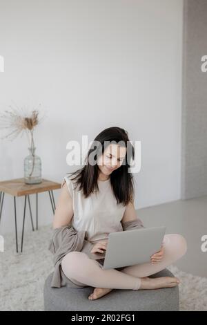 Giovane donna seduta comodamente con un computer portatile su uno sgabello nel soggiorno Foto Stock