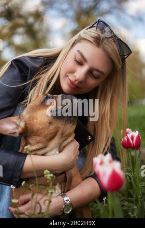 Donna amante del cane che abbraccia il cucciolo di bulldog francese. Padrone di animali domestici amorevole che gioca con il giovane cane marrone nel campo dei fiori in primavera Foto Stock