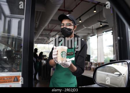 I lavoratori Starbucks danno ordini con espressioni facciali felici indossando una maschera facciale al drive-through. Caffè mocha bianco ghiacciato. Foto Stock