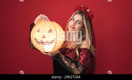 Giovane donna bionda che indossa il costume katrina tenendo la lampada di zucca sopra lo sfondo rosso isolato Foto Stock