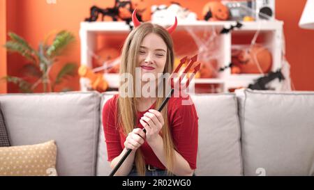 Giovane donna bionda sorridente indossando il costume diavolo seduto sul divano a casa Foto Stock