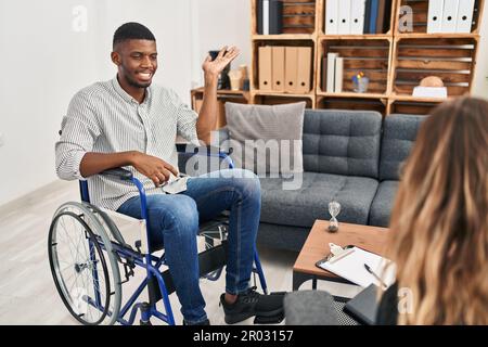 Uomo afroamericano che fa terapia seduto su sedia a rotelle sorridendo allegro presentazione e indicando con palmo di mano guardando la macchina fotografica. Foto Stock