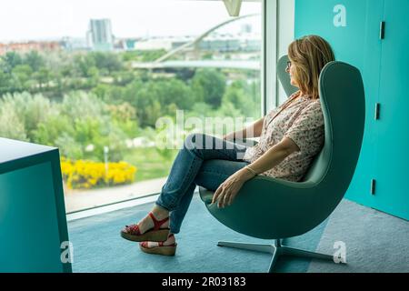 Donna seduta in una poltrona rilassata e contemplando il paesaggio da una grande finestra della stanza. Foto Stock