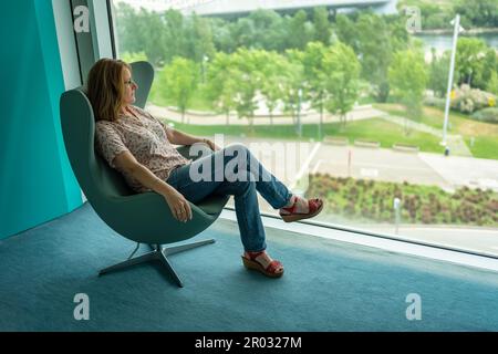 Donna seduta in una poltrona rilassata e contemplando il paesaggio da una grande finestra della stanza. Foto Stock