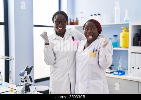 Due donne africane che lavorano presso il laboratorio di scienziato urlano orgogliose, celebrando la vittoria e il successo molto eccitate con le armi sollevate Foto Stock