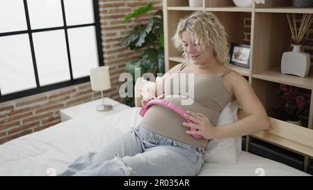 Giovane donna incinta sorridente sicura di mettere le cuffie sul ventre in camera da letto Foto Stock