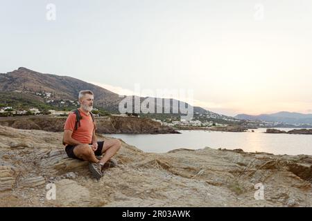 Uomo adulto con una barba bianca seduta su alcune rocce meditando dal mare con gli occhi chiusi. Foto Stock