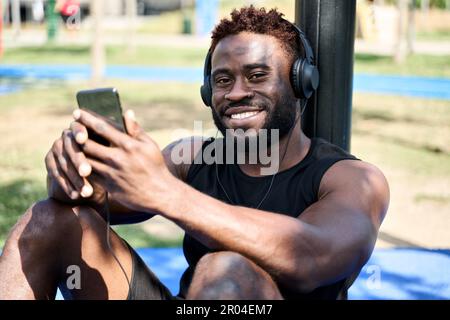 Felice giovane uomo nero seduto nel parco allenamento utilizzando le app mobili ascoltando musica. Foto Stock