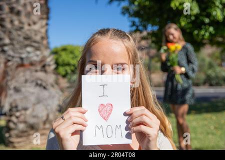 La figlia sta tenendo la scheda che dice 'i Love mom' con la sua madre dietro con i fiori ricevuti come regalo Foto Stock