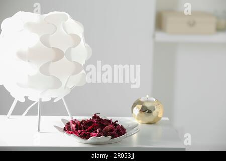 Piatto con potpourri aromatici, lampada e candela a fuoco su tavolo bianco in camera Foto Stock