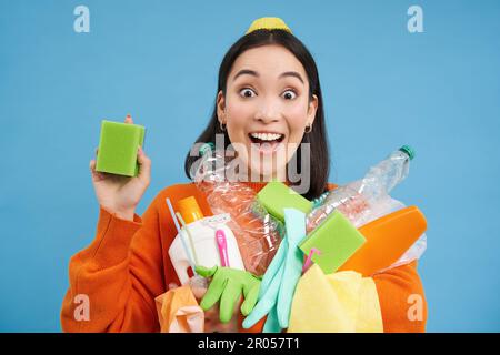 Ragazza asiatica entusiasta mostrando spugne di pulizia, tenendo bottiglie di plastica vuote, rifiuti per il riciclaggio, la cernita della sua spazzatura di casa, sfondo blu Foto Stock