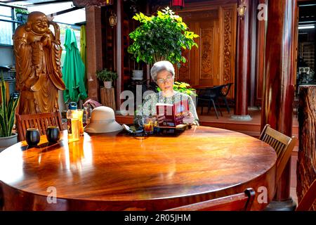 Una donna anziana legge un libro al caffè Nha Go nella città di PLEIKU, Vietnam Foto Stock