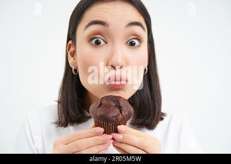 Primo piano di bella donna asiatica sorridente, tenendo cupcake al cioccolato vicino alla bocca, avendo morso, ama mangiare pasticceria, sfondo bianco Foto Stock