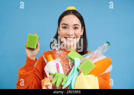 Ragazza asiatica entusiasta mostrando spugne di pulizia, tenendo bottiglie di plastica vuote, rifiuti per il riciclaggio, la cernita della sua spazzatura di casa, sfondo blu Foto Stock