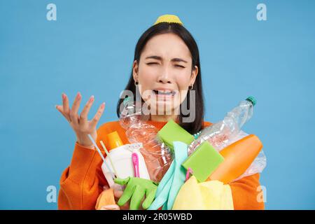 Frustrata donna asiatica che piange, tenendo carichi di bottiglie di plastica vuote e rifiuti per il riciclaggio, cercando stressato, sfondo blu Foto Stock