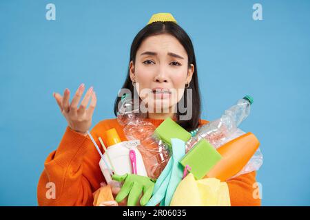 Frustrata donna asiatica che piange, tenendo carichi di bottiglie di plastica vuote e rifiuti per il riciclaggio, cercando stressato, sfondo blu Foto Stock