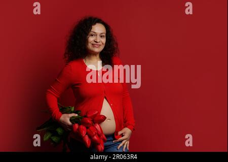 Donna incinta multietnica sorride guardando la macchina fotografica, tiene un bouquet di tulipani rossi su sfondo di colore isolato Foto Stock