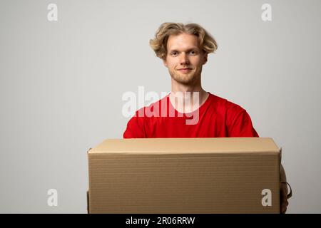Servizio di consegna. Corriere sorridente giovane che tiene la scatola di cartone. Uomo di consegna giovane felice in t-shirt rossa in piedi con pacco isolato su bianco Foto Stock