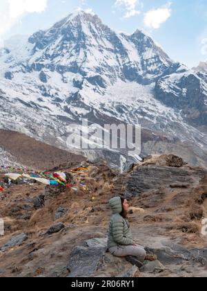 Vista verticale di una donna seduta nel campo di base Annapurna facendo una meditazione per ringraziare gli spiriti della natura dopo tutto lo sforzo che ha preso per arrivare. Foto Stock