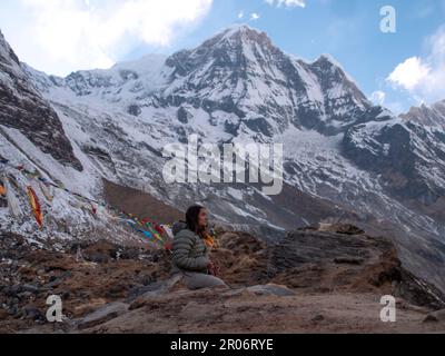 Donna zaino in spalla seduto nel campo di base Annapurna facendo una meditazione per ringraziare gli spiriti della natura dopo tutto lo sforzo che ha preso per arrivare lì. Foto Stock