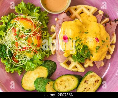 Uova Benedict (uova in camicia) su prosciutto alla griglia con salsa olandese su cialde belghe con zucchine alla griglia Foto Stock