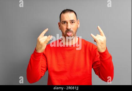 Latino uomo con una barba nel suo 40s indossando un maglione rosso facendo il gesto rock mentre si stacca la lingua in un atteggiamento pazzo, isolato sul giallo Foto Stock